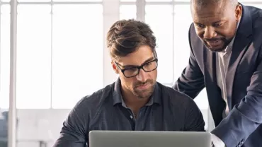 Dos hombres trabajando juntos frente a una laptop en una oficina luminosa.